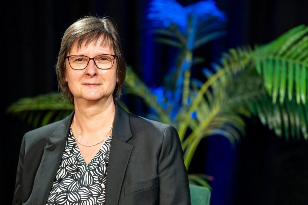 Petra Borchardt in einem Portraitfoto vor blauem Hintergrund und einer Pflanze.